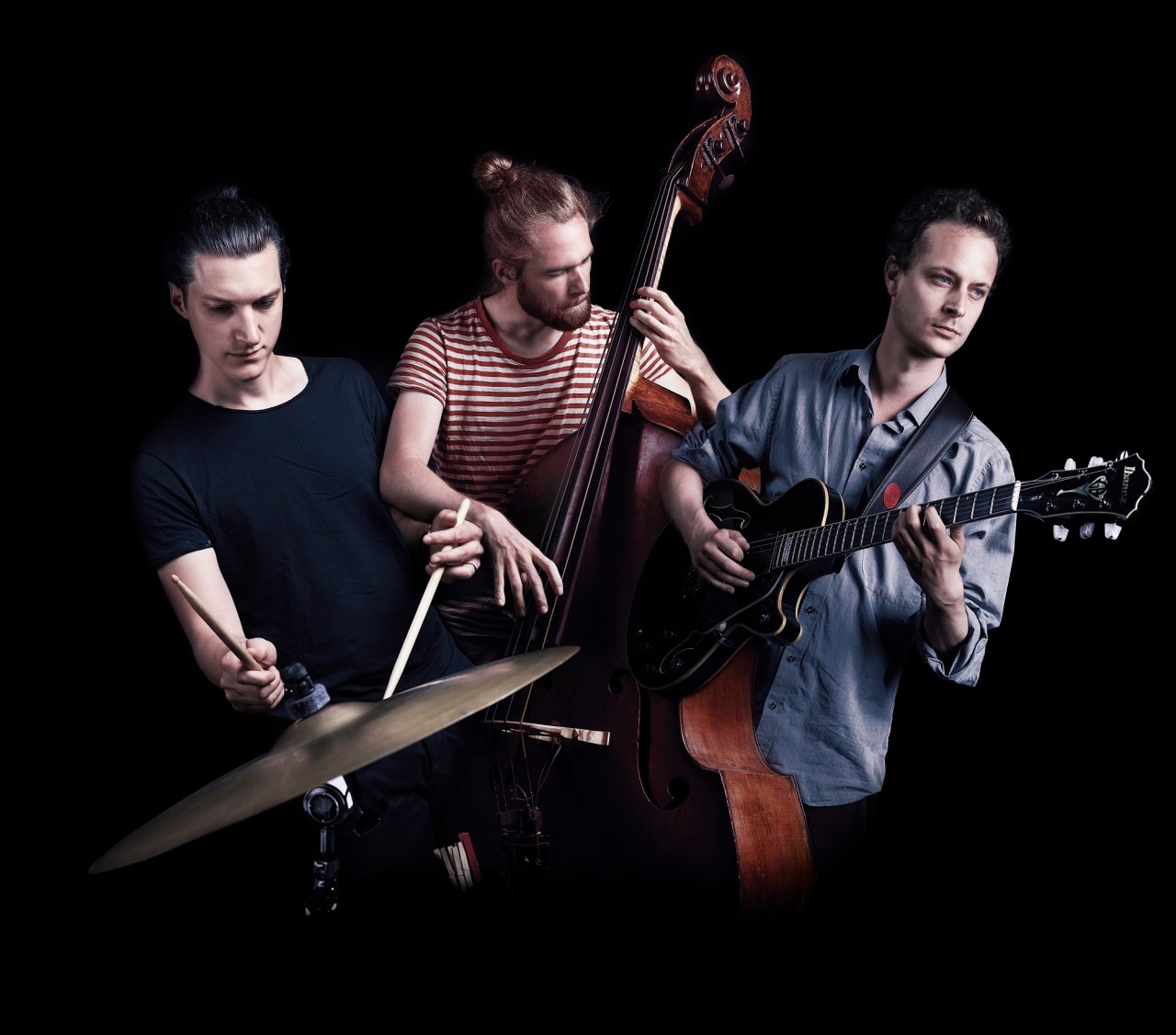 Drei Musiker mit Kontrabass, Gitarre und Schlagzeug befinden sich auf einer dunklen Bühne.