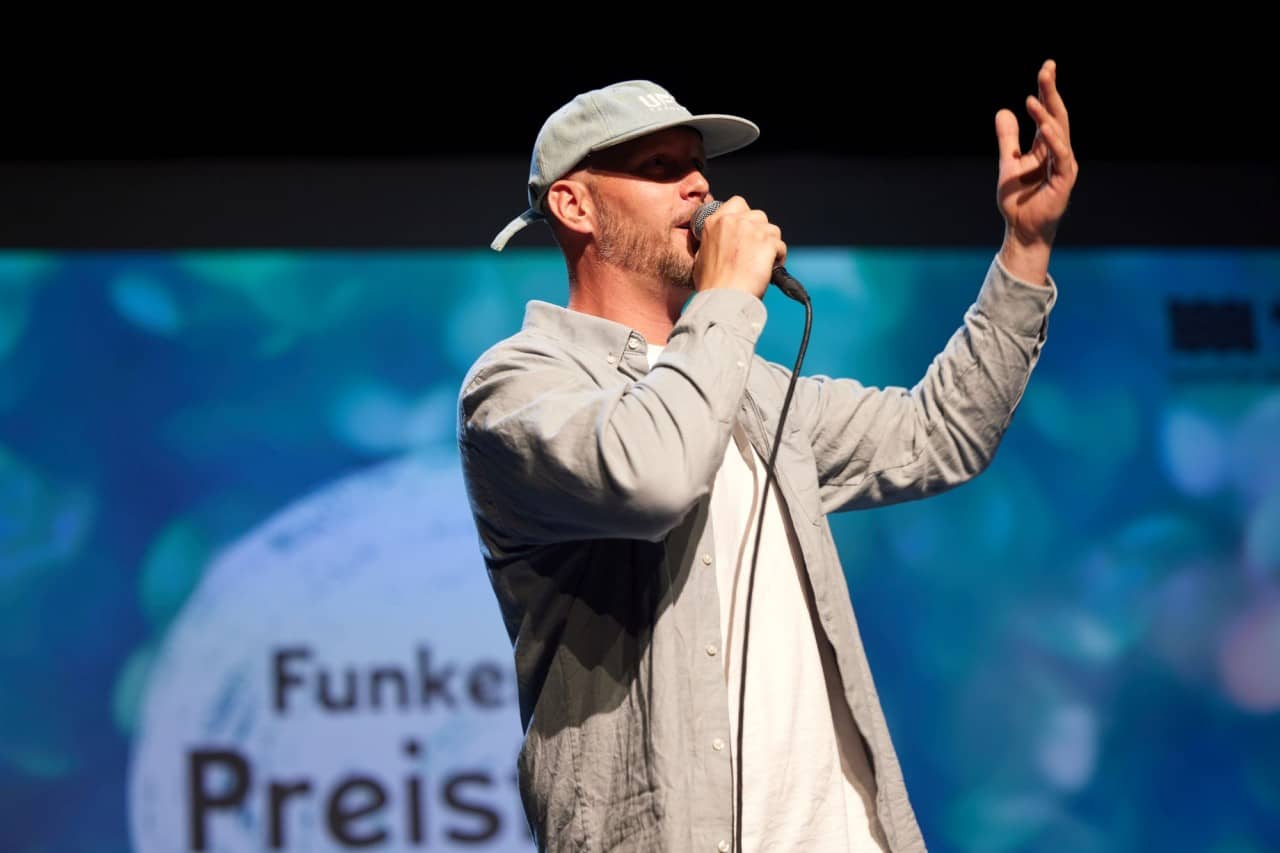 Mann mit grauem Hemd und Mütze singt in ein Mikrofon.