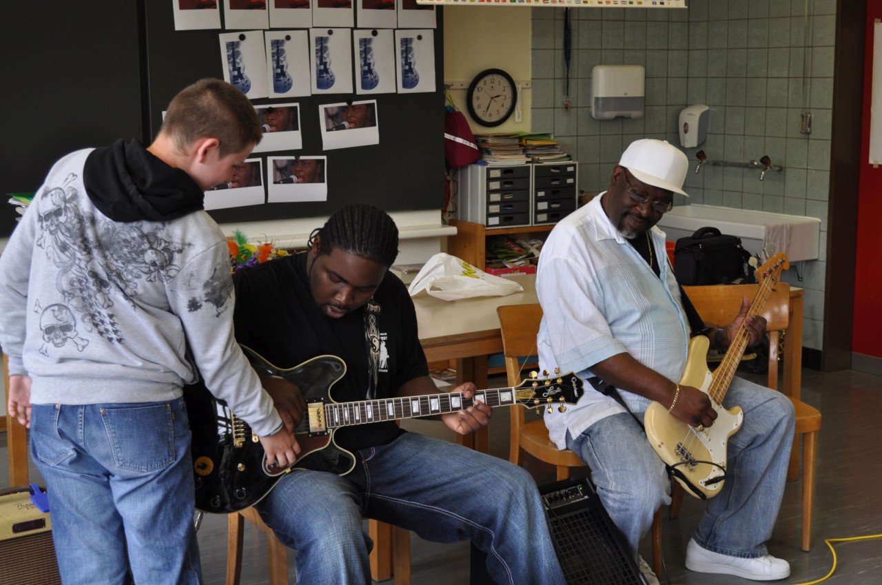 Zwei Männer sind in einem Schulzimmer und zeigen ihre Gitarren einem Schüler.