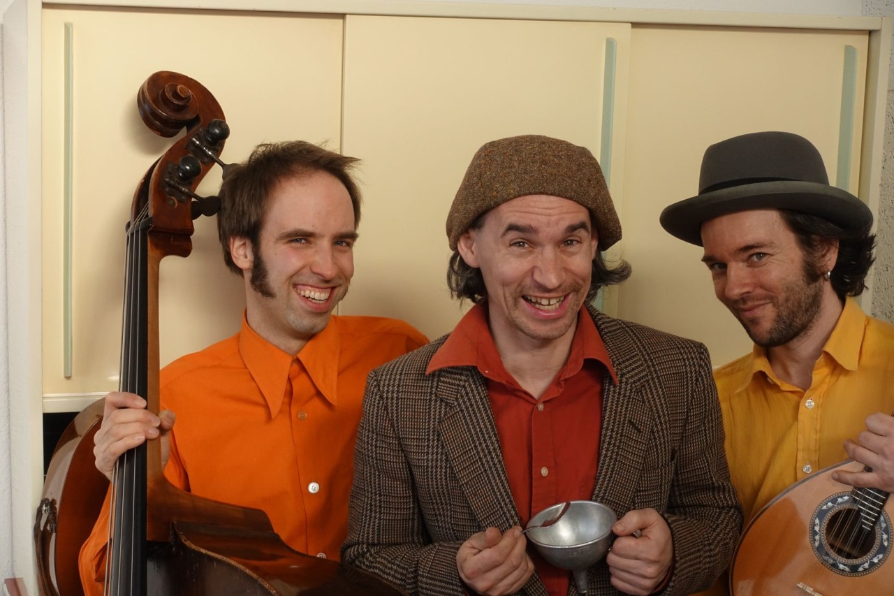 Drei Männer lächeln in die Kammera. Einer hält einen Kontrabass, ein Mann mit Hut hält eine Tasse und einen Löffel und ein weiterer Mann hält ein Saiteninstrument. 