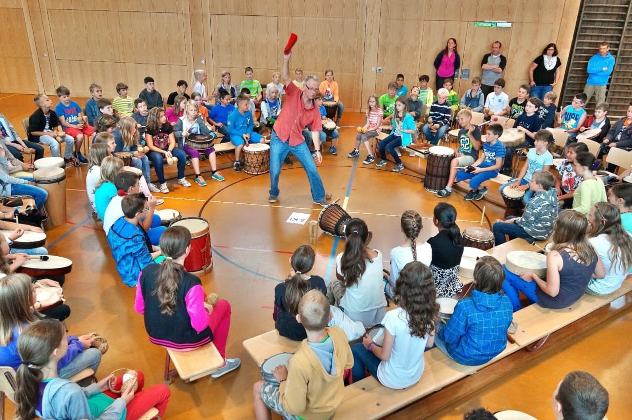 Schülerinnen und Schüler sitzen im Kreis und spielen auf Trommeln. In der Mitte steht MArhias Schiesser und gibt ein Zeichen. 