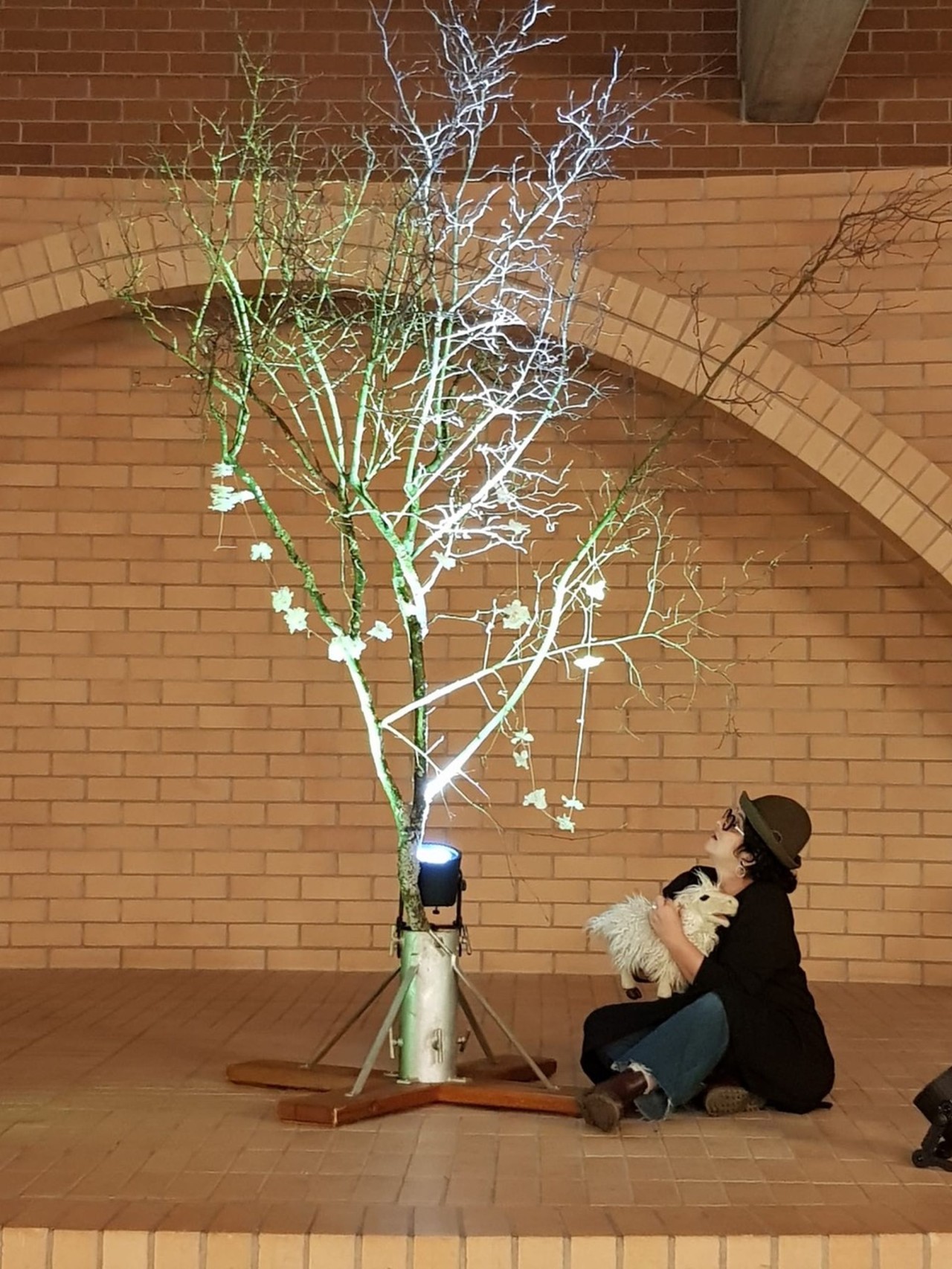 Person sitzt vor einer Backsteinwand neben einem angestrahlten kahlen Baum mit einzelnen Blumenformen. Person hält ein weisses Stofftier.
