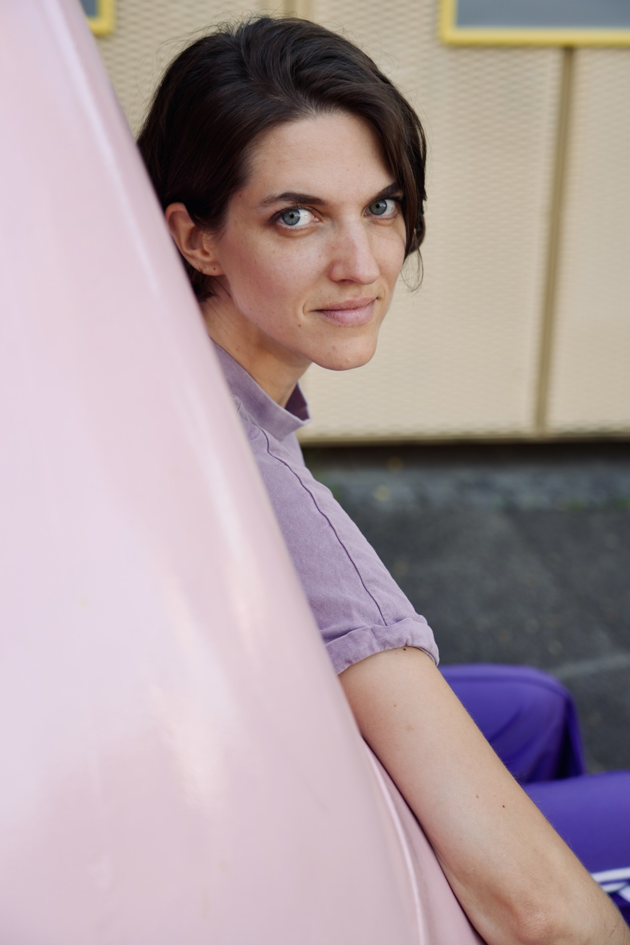 Eine Person mit kurzen dunkelbraunen Haaren in lila Kleidung lehnt an einem grossen rosa Gegenstand, Blick in die Kamera, Hintergrund mit beige Wand und Asphalt.