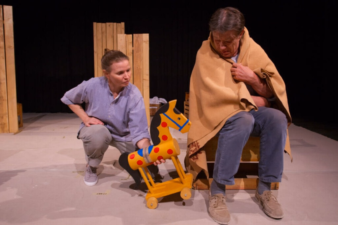 Zwei Personen sind mit einem Holzpferd auf einer Bühne. Sie interagieren mit dem Pferd.