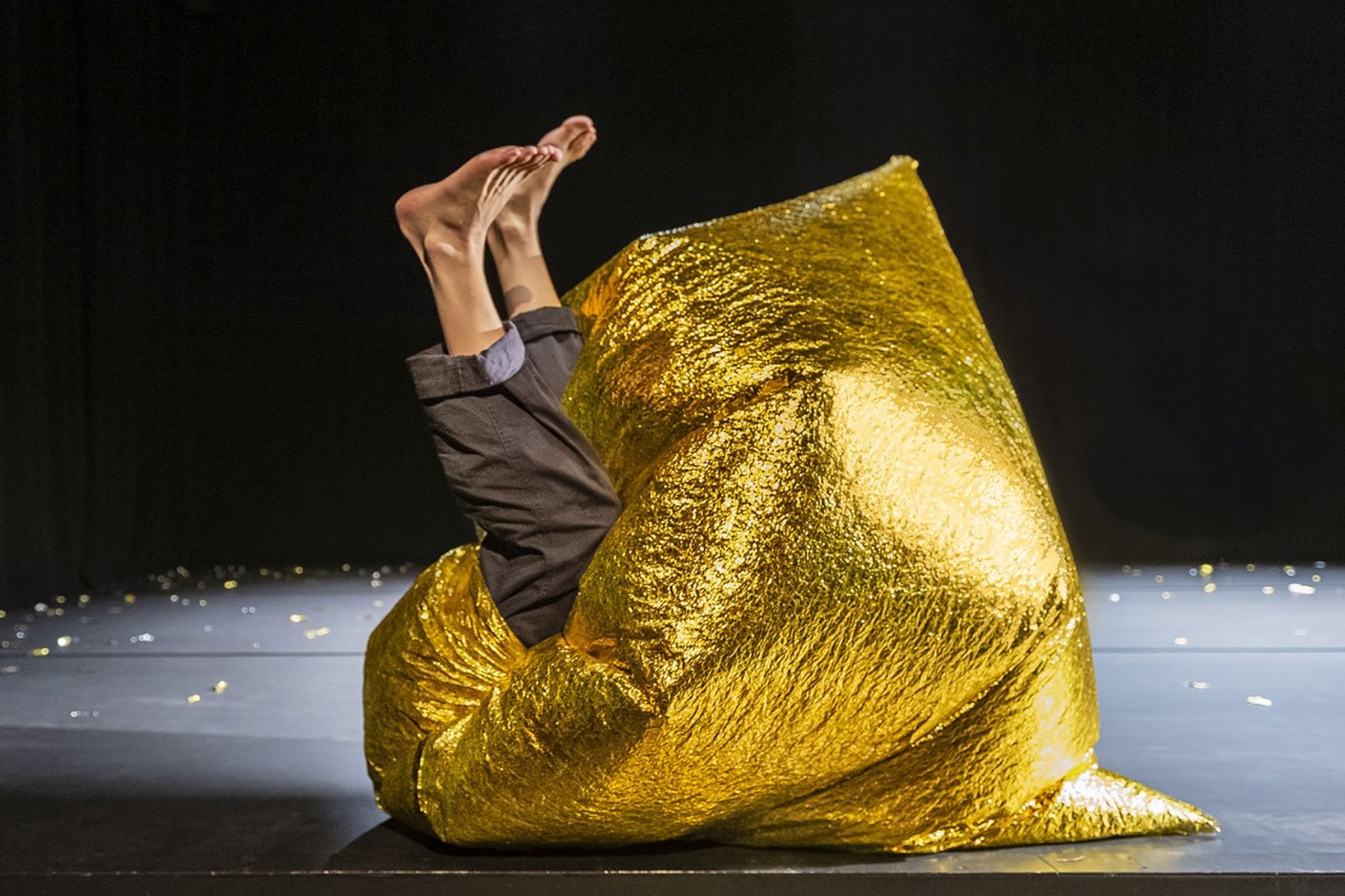 Beine einer Person ragen aus einem grossen goldenen Stoffbeutel auf einer Theaterbühne in die Höhe.