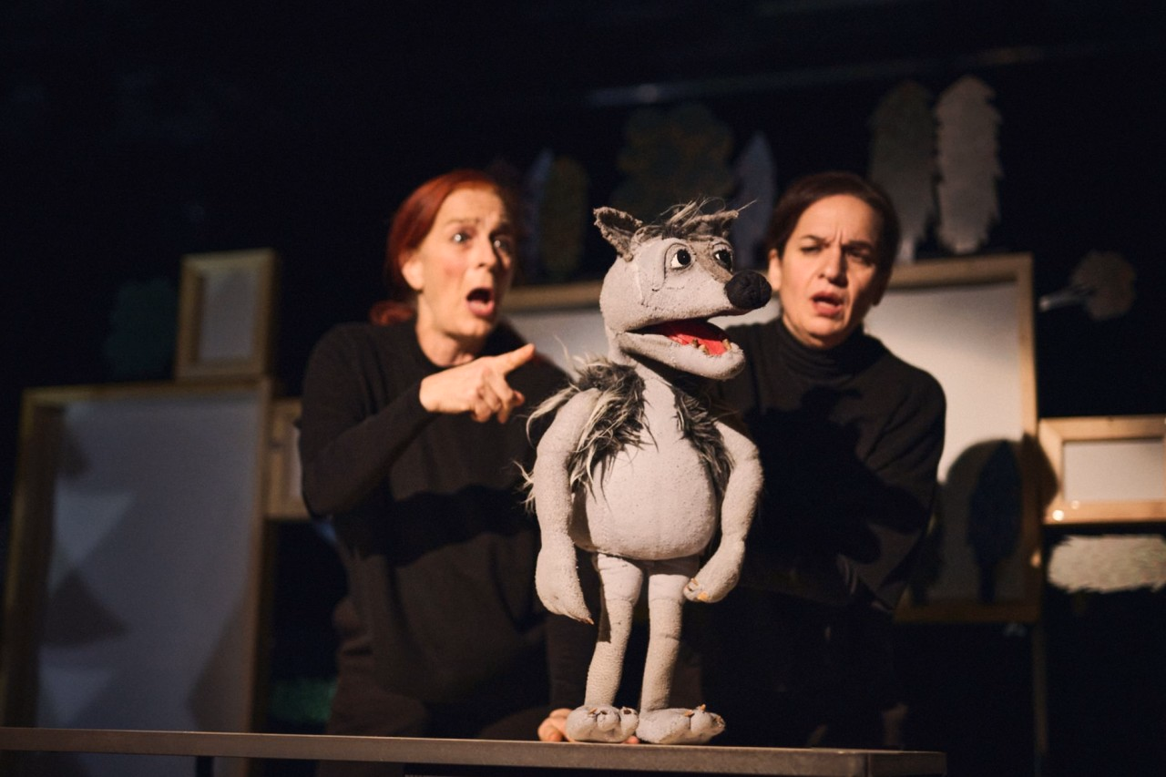 Zwei Personen führen auf einer Bühne eine grosse graue Handpuppe in Wolfsgestalt.