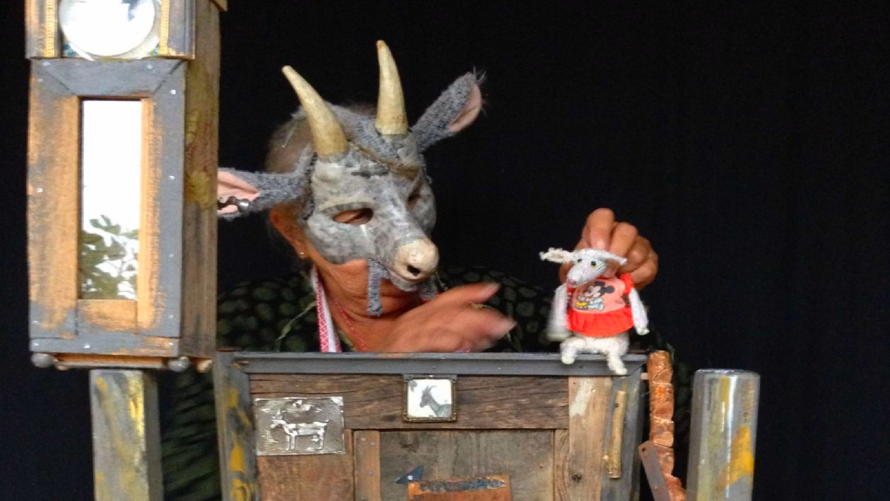 Eine Figurenspielerin trägt eine Ziegenmaske. Sie hält eine kleine Geissenfigur.