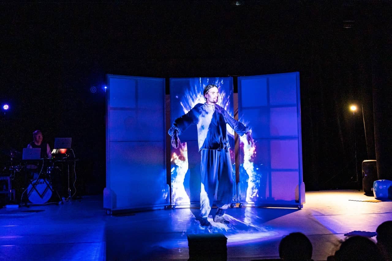 Ein Mann steht auf einer Bühne, er wird mit einem blauem Licht beleuchtet. 