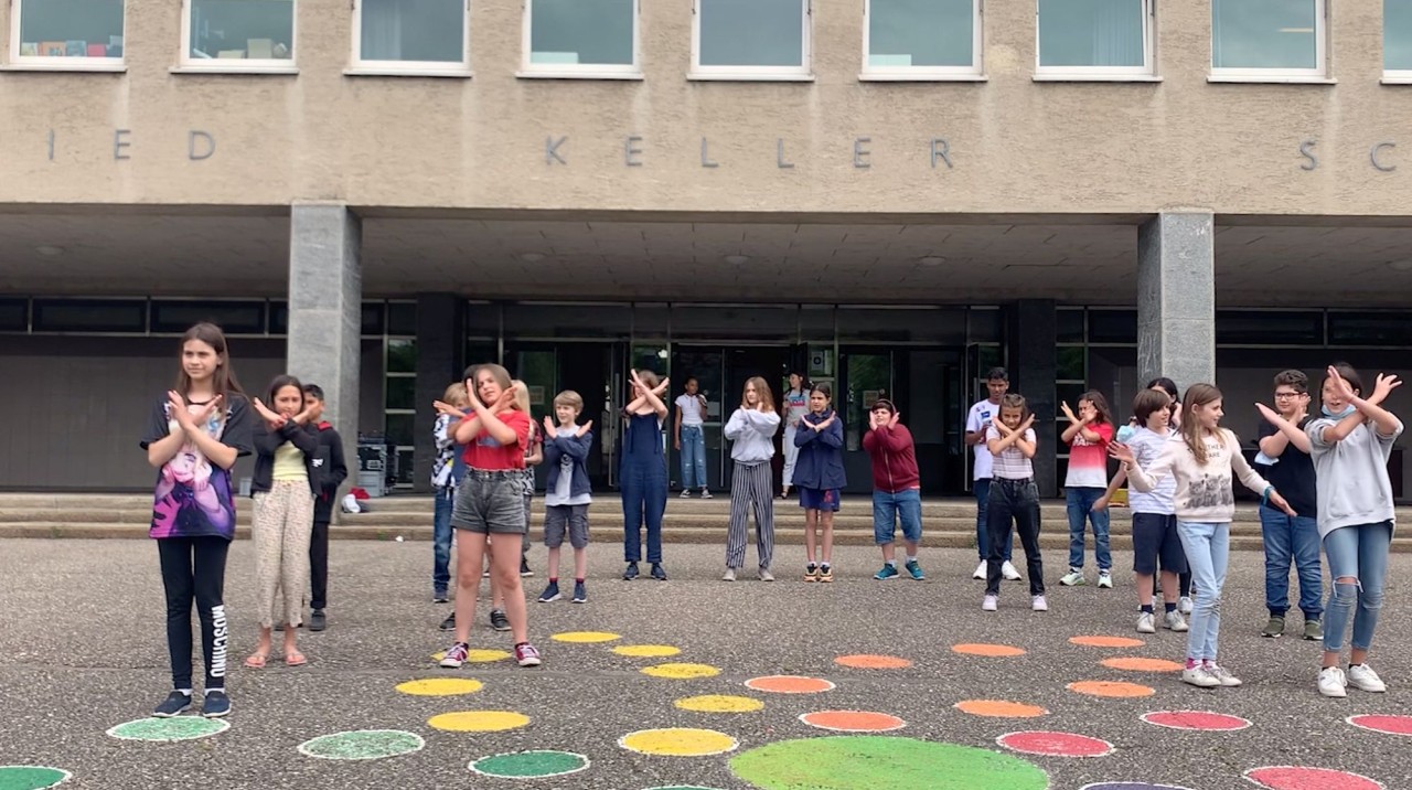 Schülerinnen und Schüler stehen vor dem Schulhaus auf bunten Kreisen und kreuzen die Arme.