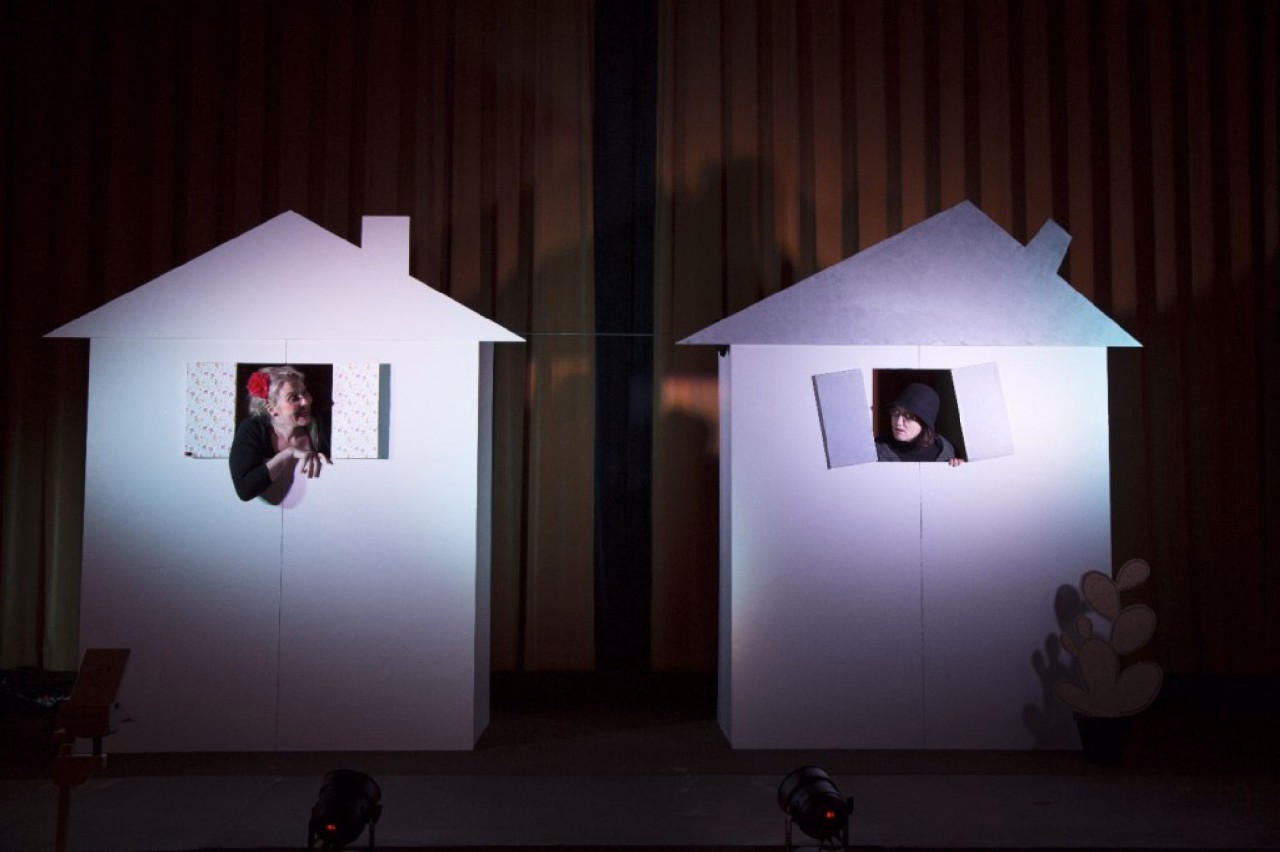 Auf der Bühne befinden sich zwei Häuser. Zwei Frauen unterhalten sich durch die Fenster.