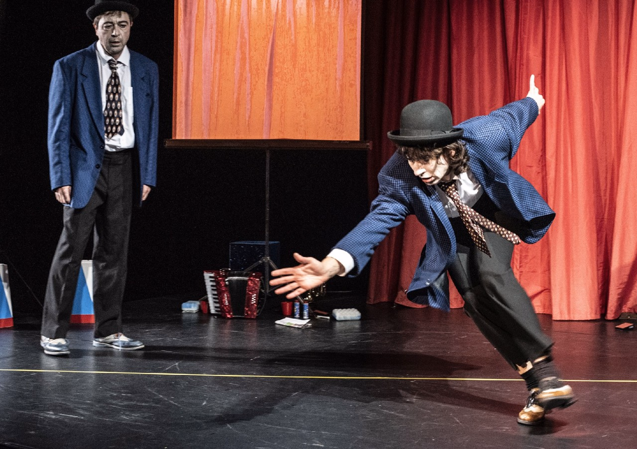 Zwei Clowns befinden sich auf einer Bühne. Der eine Clown stolpert nach vorne.