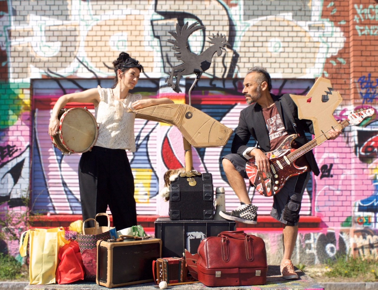 Zwei Personen befinden sich vor einem besprayten Gebäude, sie halten Instrumente und schauen sich an.