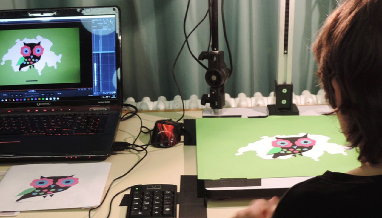 Kind arbeitet am Trickfilmset. Auf dem Tisch liegen Laptop und die Figur einer bunten Eule vor grünem Hintergrund.
