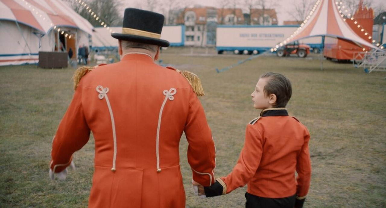 Zirkusdirektor und Kind in roten Uniformen gehen Hand in Hand über einen Platz zwischen Zirkuszelten.