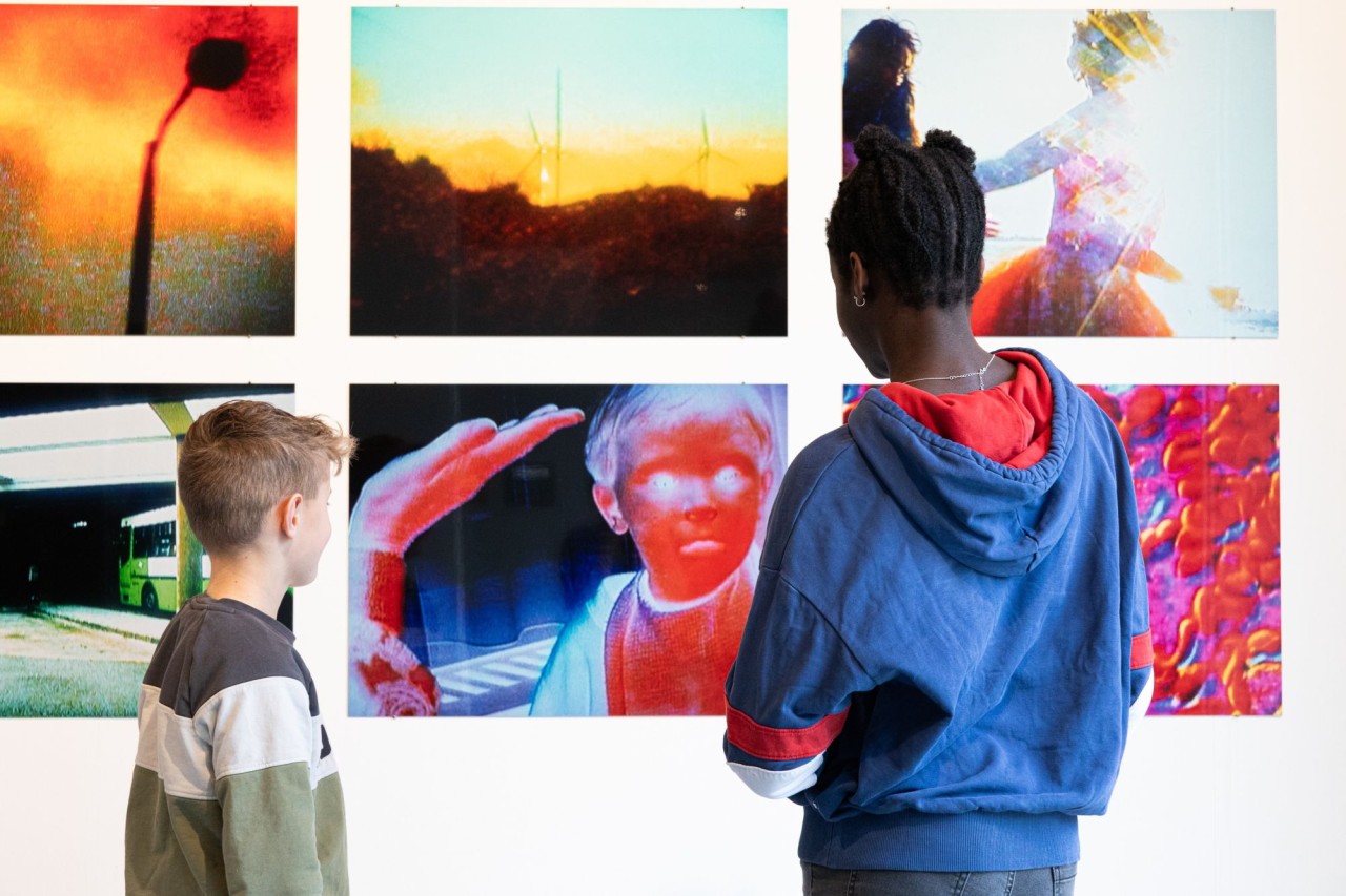 Zwei Schülerinnen und Schüler stehen vor einer Wand, an der farbige Fotografien und Plakate hängen.