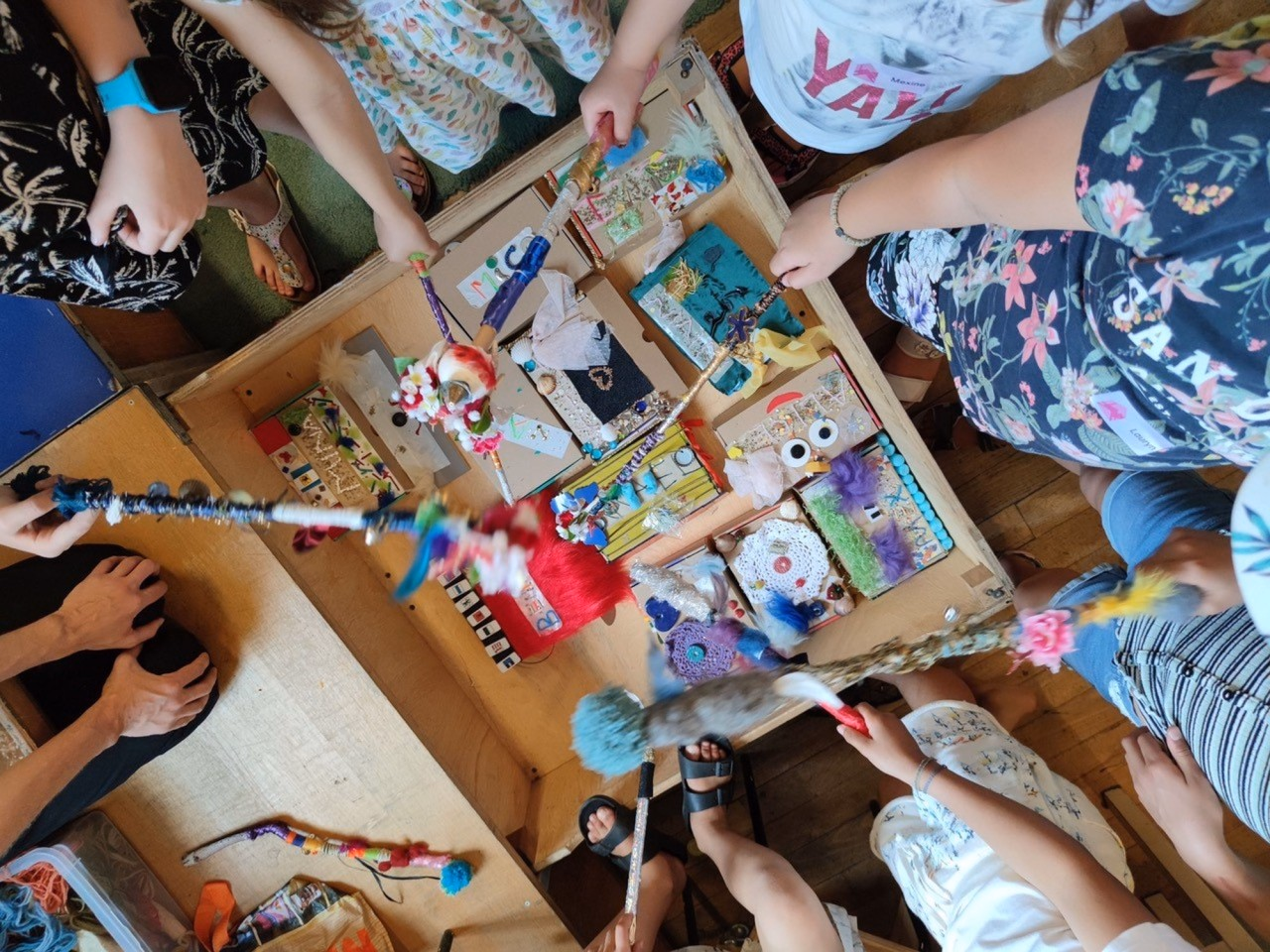 Kinder stehen um einen Holzkasten mit bunt gestalteten kleinen Objekten und halten verzierte Stäbe in den Händen.