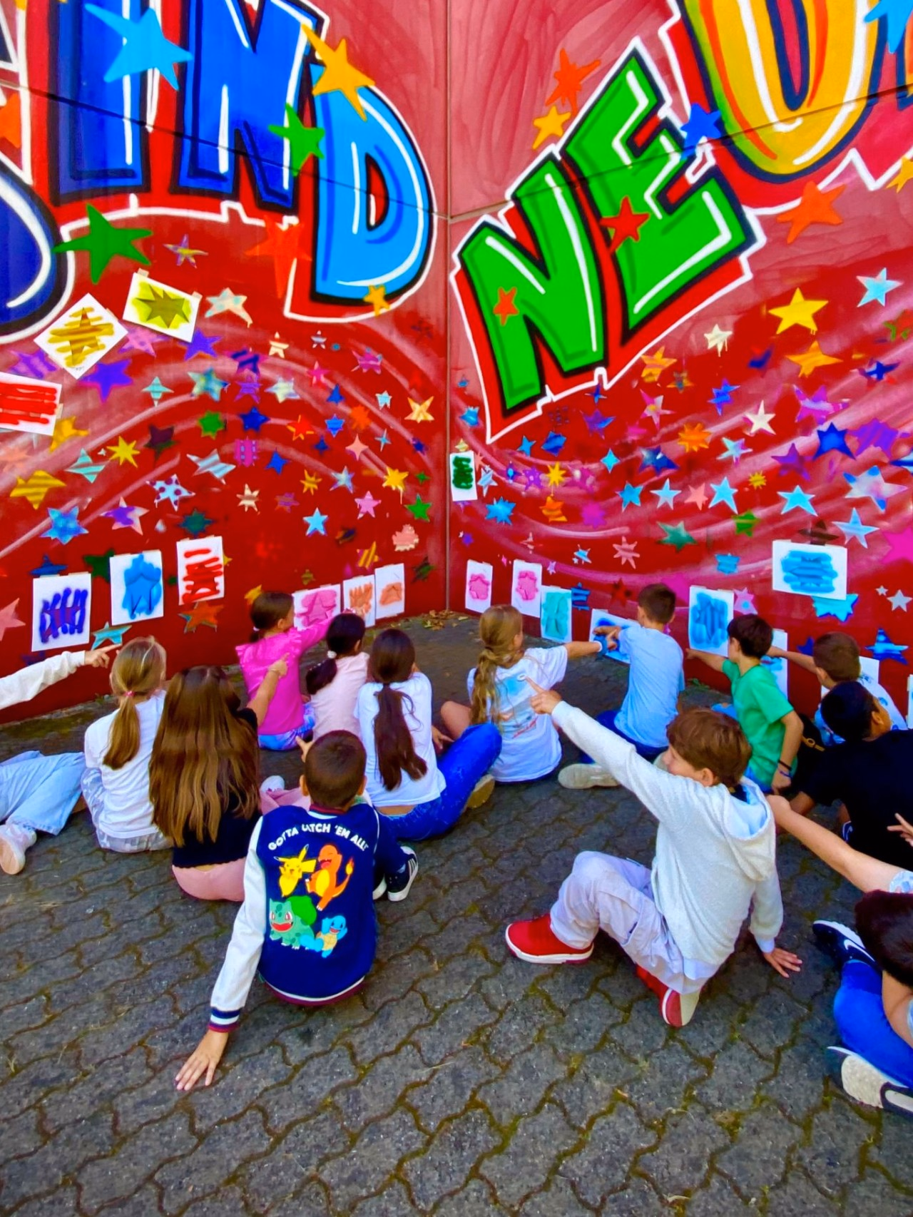 Kinder sitzen vor einer roten Wand mit buntem Graffiti-Schriftzug und zeigen auf ihre aufgehängten Bilder.