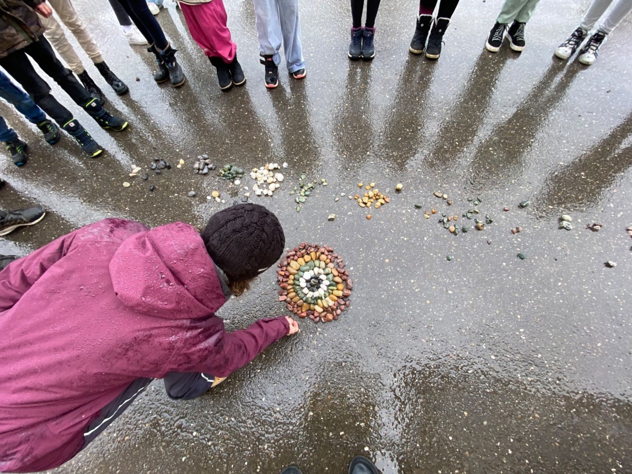 Gruppe von Kindern steht draussen im Regen in einem Kreis. Eine Person legt Steine und andere Naturalien zu einem Mandala.
