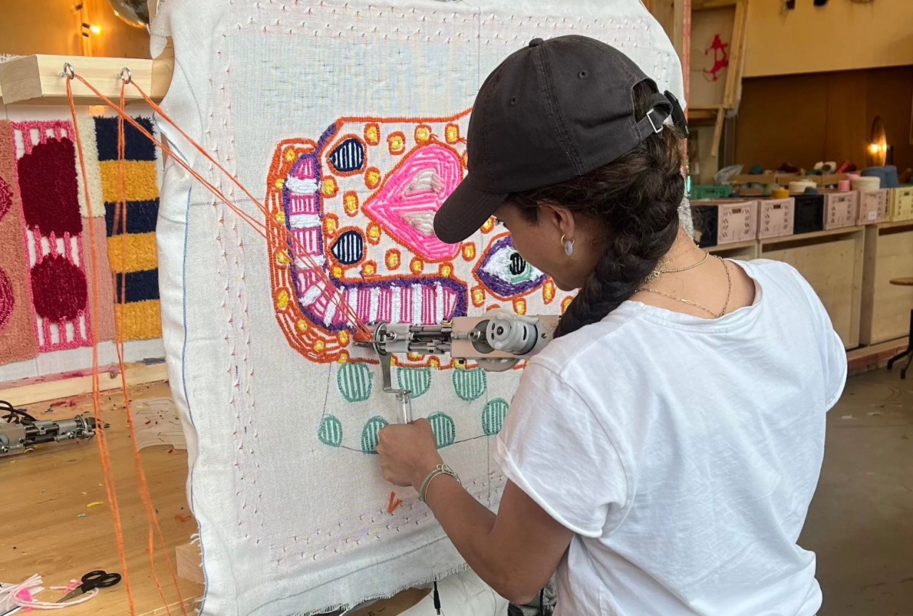 Person arbeitet in einer Werkstatt mit einer Tufting-Maschine an einem farbigen Textilbild auf einem Rahmen.