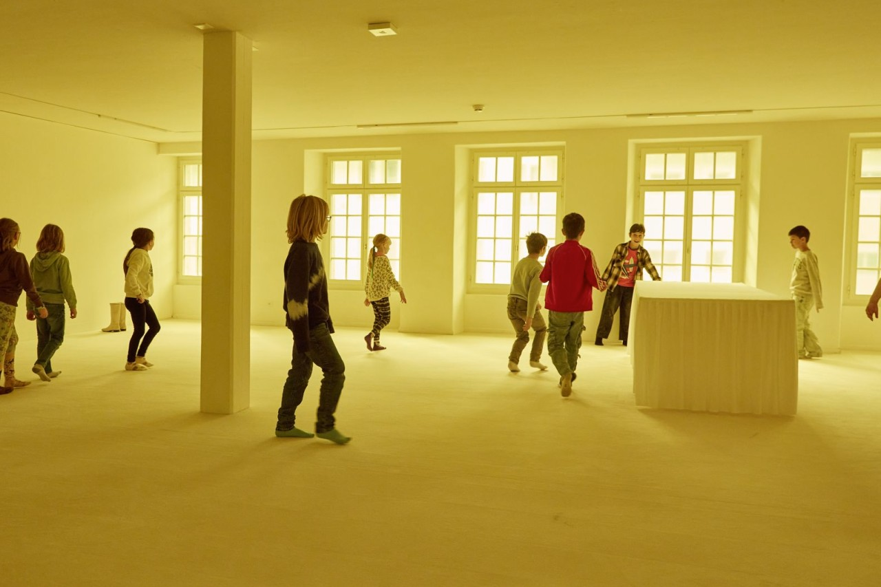 Kinder bewegen sich in einem grossen, hellgelben Raum mit vielen Fenstern und einem weissen Podest.