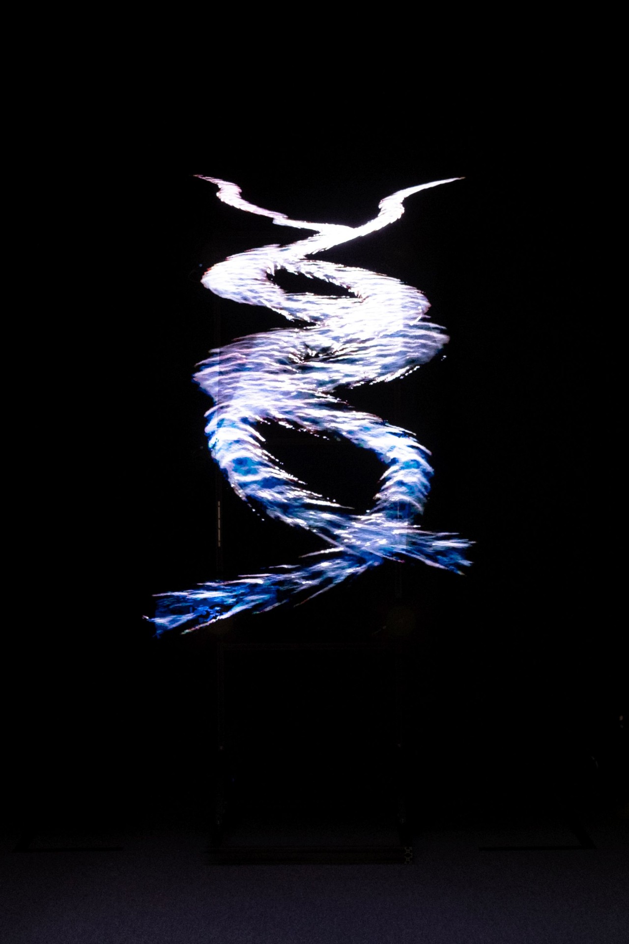 Hologramm, vor dunklem Hintergrund, bestehend aus einem geschwungenen, spiralförmigen Lichtstreifen in blau-weissen Tönen.