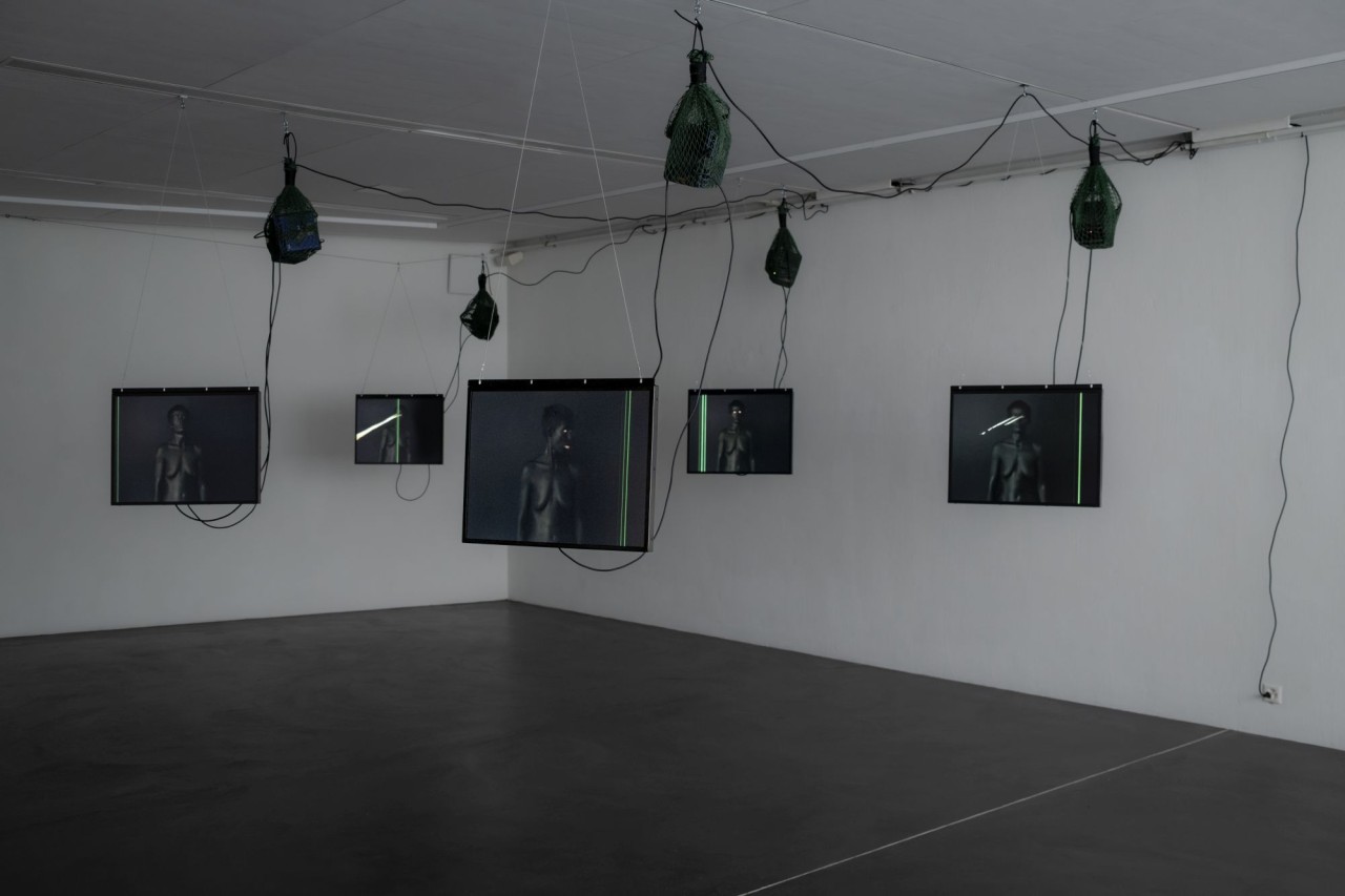 Mehrere Bildschirme hängen an einer Wand in einem dunklen Raum. Auf den Bildschirmen sind schwarz-weisse Filme mit Personen und Neonstreifen zu sehen. Flaschen sind an Kabeln befestigt.