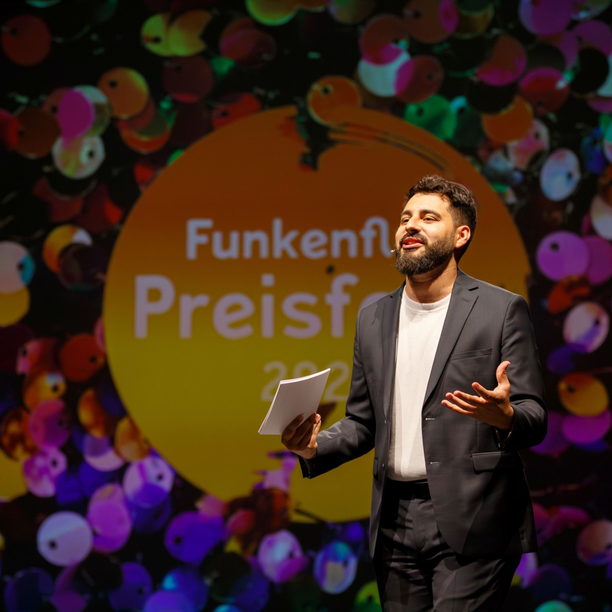 Ein Mann in dunkelgrauem Anzug steht vor einer bunten, kreisförmigen Kulisse mit der Aufschrift "Funkenflug Preisfeier 2026" und hält ein Blatt Papier in der Hand.