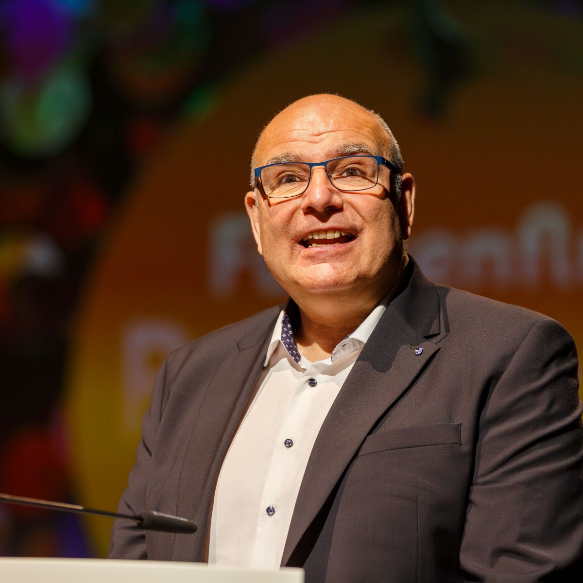 Ein Mann steht hinter einem Rednerpult, trägt ein dunkles Sakko und ein weißes Hemd. Der Hintergrund zeigt bunte Lichter und einen orangefarbenen Kreis mit teilweise lesbarem Text.
