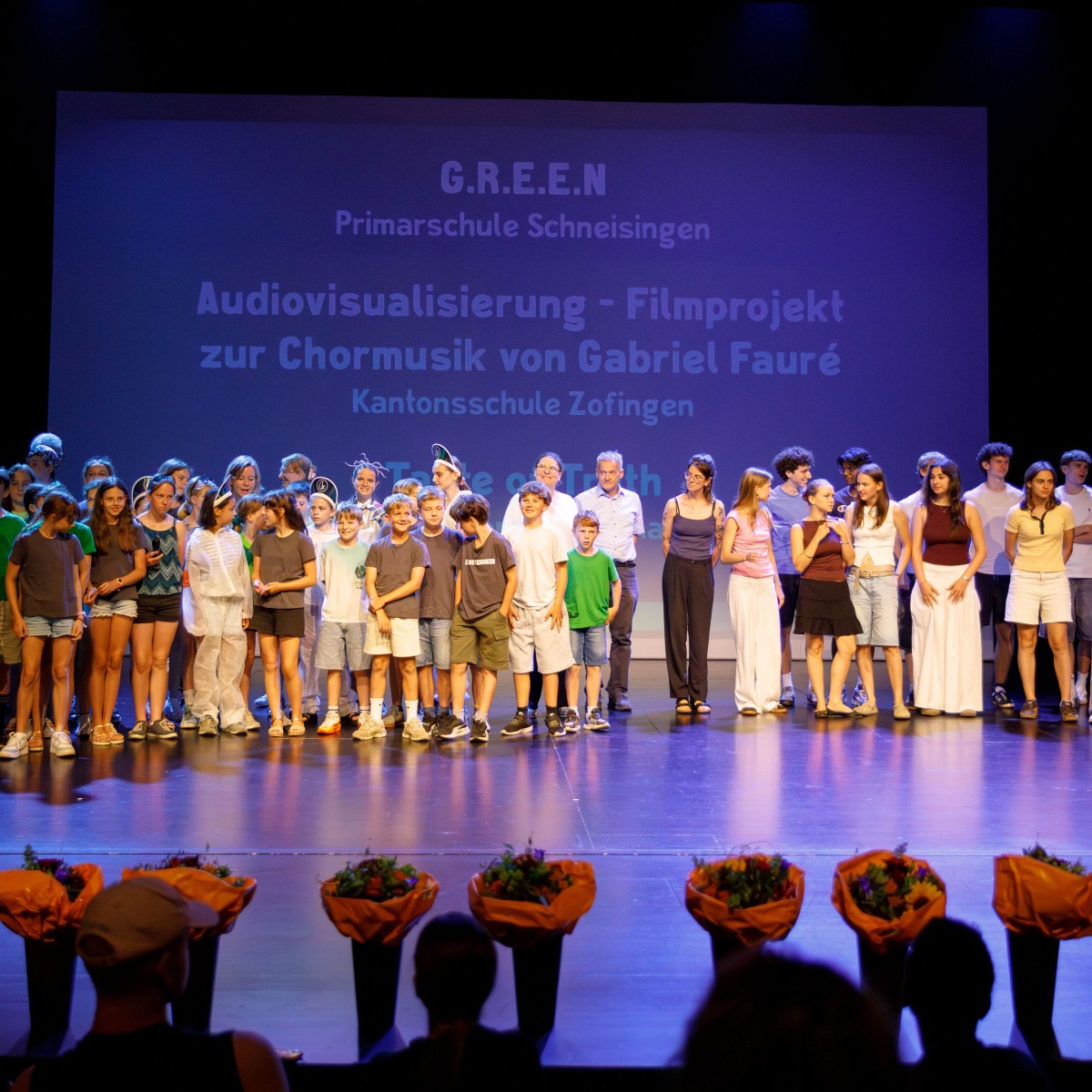 Eine grosse Gruppe von Schülerinnen, Schülern und Erwachsenen steht auf einer Bühne vor blauem Hintergrund.