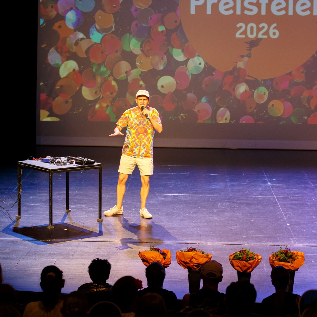 Ein Mann in bunter Kleidung und Mütze steht auf einer Bühne vor einem DJ-Pult. Im Hintergrund eine Projektion mit dem Text "Preisfeier 2026". Im Vordergrund sind Zuschauende mit Blumensträussen sichtbar.