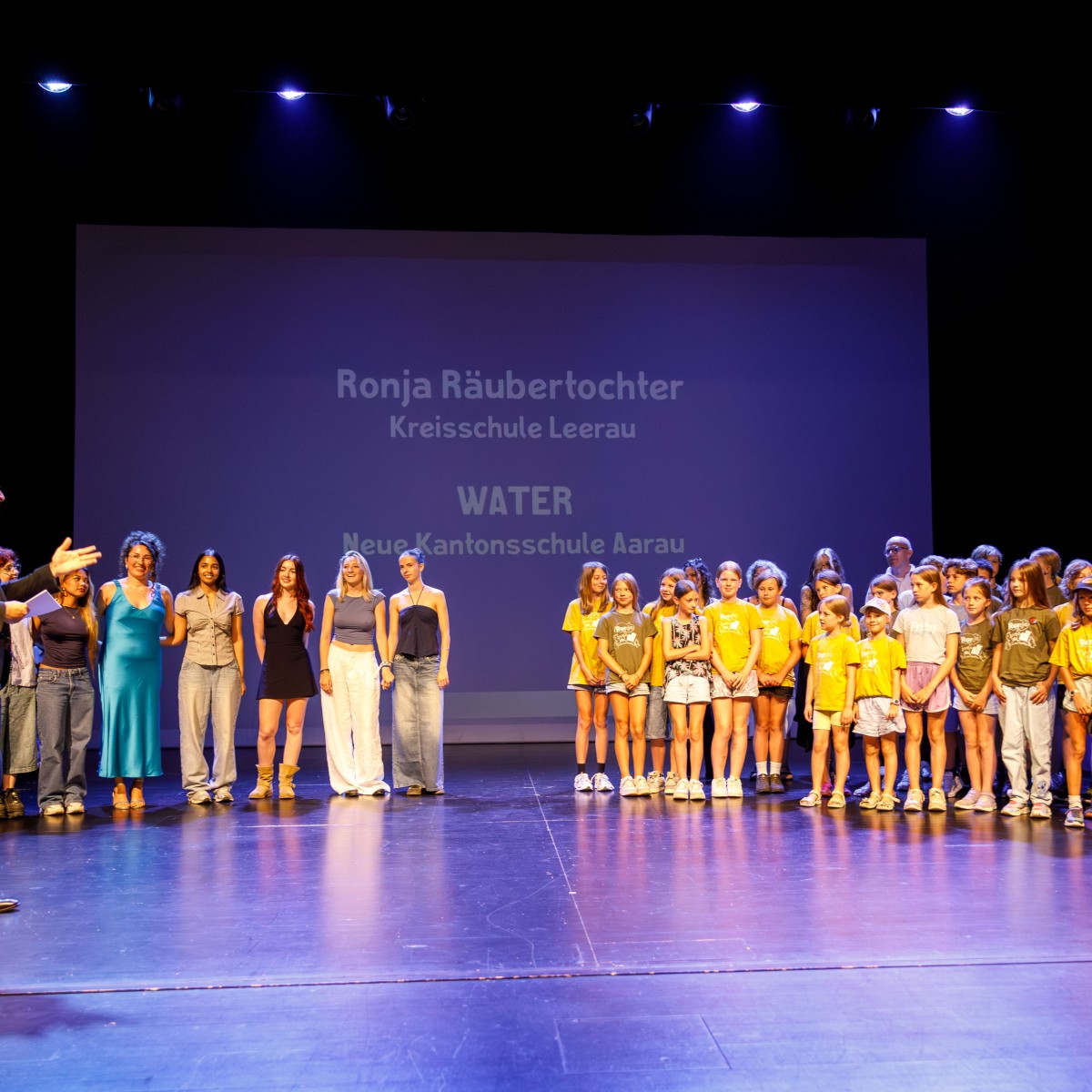 Eine grosse Gruppe von Kindern und Erwachsenen steht auf einer Bühne. Im Hintergrund ist der Schriftzug "Ronja Räubertochter Kreisschule Leerau" und "WATER Neue Kantonsschule Aarau" auf einer Leinwand sichtbar.