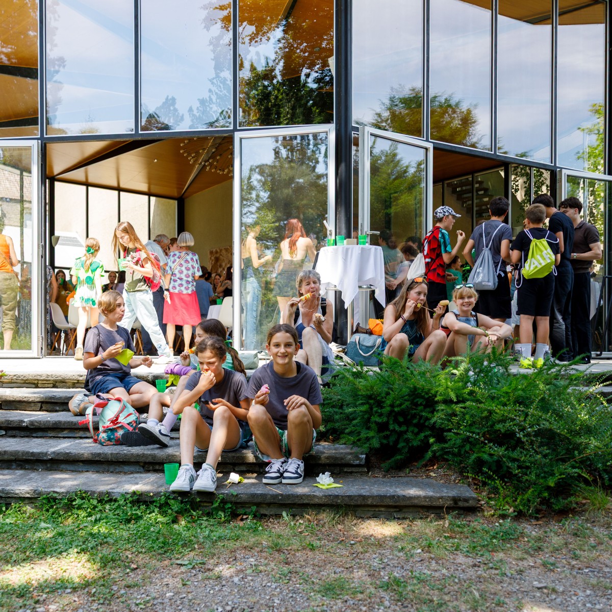 Kinder und Jugendliche sitzen auf Steinstufen vor einem gläsernen Gebäudeeingang, einige essen und trinken, während andere stehen.
