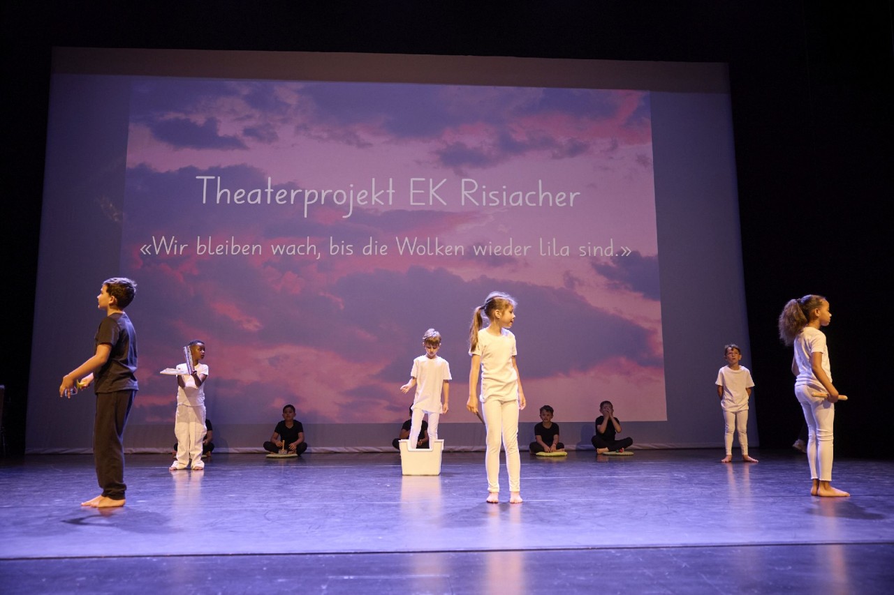 Schülerinnen und Schüler in schwarzer und weisser Kleidung tanzen und performen auf einer Bühne. Im Hintergrund eine Präsentation mit Titel des Projekts in lila Farben.