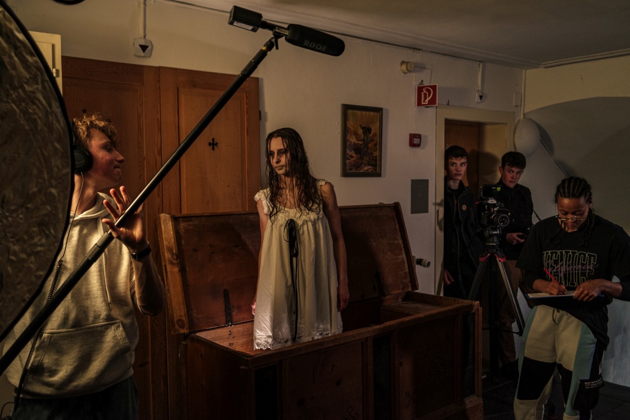 Foto von jungen Menschen bei einem Filmdreh. Junge Frau steht in unheimlichem Kostüm in einer Holztruhe. Über ihr ein Mikrofon. Im Hintergrund weitere Personen mit technischer Ausrüstung.