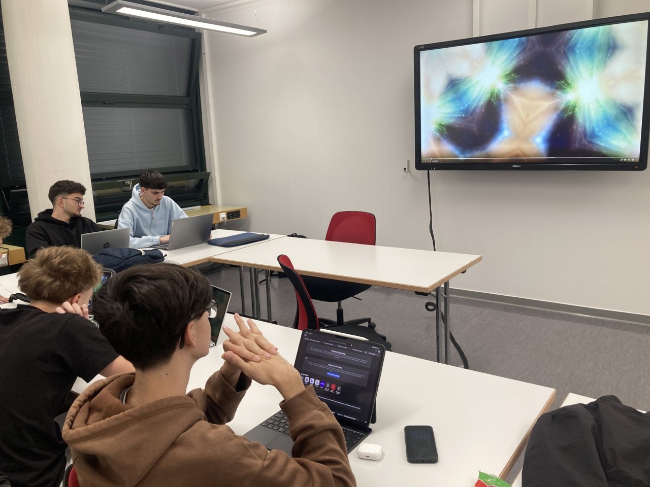 Mehrere junge Menschen sitzen an weissen Tischen mit Laptops in einem modernen Raum. An der Wand hängt ein Bildschirm, der eine bunte, abstrakte Animation zeigt.
