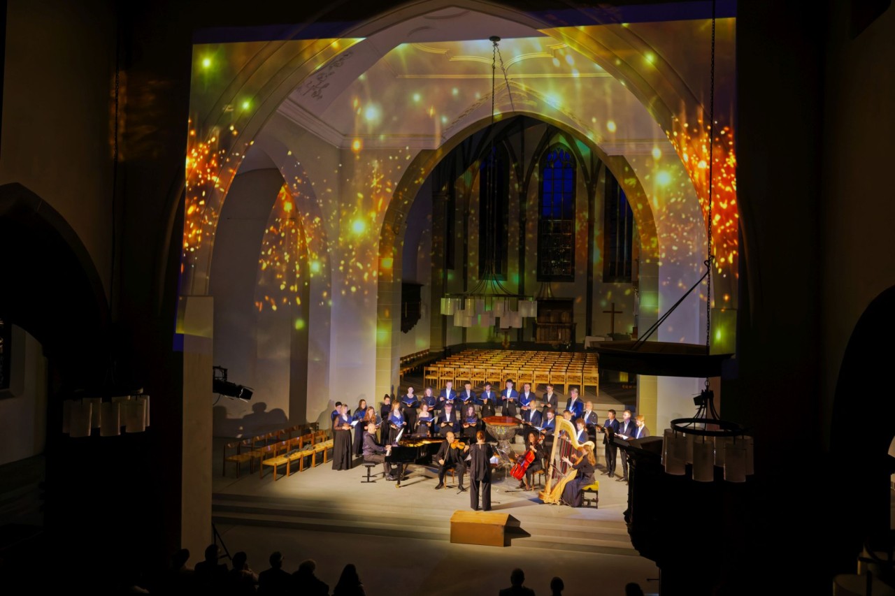 Ein Konzert in einer Kirche mit Visualisierungen an den Wänden, Chor und Orchester auf der Bühne.