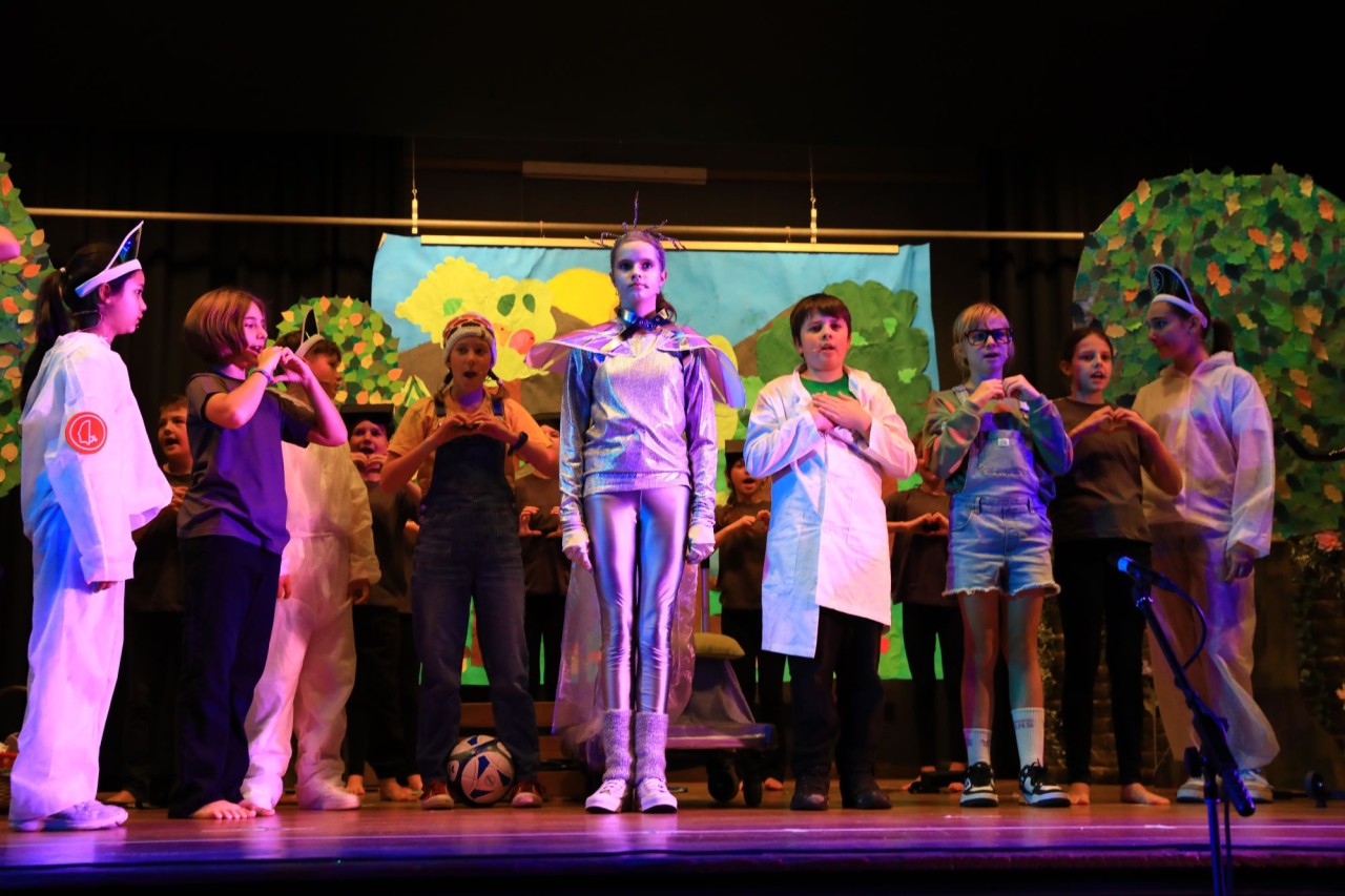 Kinder führen ein Theaterstück auf einer Bühne mit farbenfroher Kulisse auf. Verschiedene Kostüme wie Kittel, Uniformen und Tieranzüge sind zu sehen.