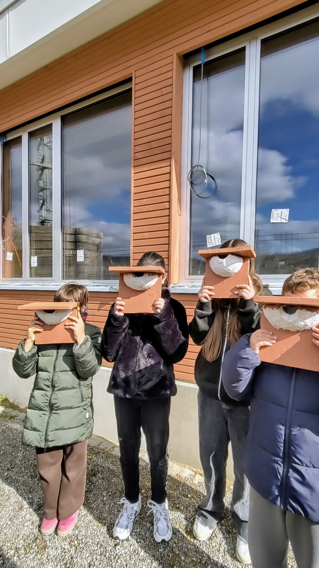 Vier Kinder stehen nebeneinander vor einem Gebäude mit brauner Fassade und Fenstern. Alle halten Masken vor ihre Gesichter.