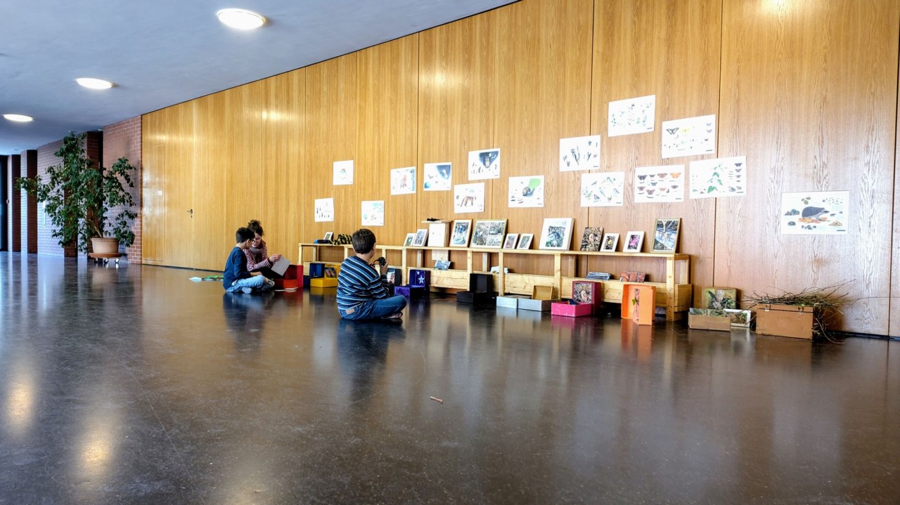 Ein Flur mit Holzwänden, an denen Zeichnungen und Poster hängen. Drei Personen sitzen auf dem Boden vor Regalen mit Büchern und Kästen. Links sind Pflanzen sichtbar.