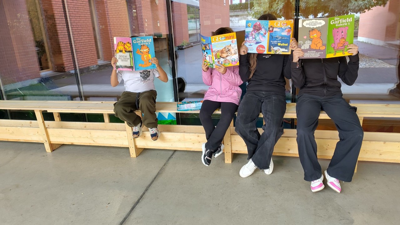 Vier Personen sitzen auf einer Holzbank und halten Comics mit Garfield-Motiven vor ihre Gesichter. Im Hintergrund sind Glasfenster und Backsteinmauern sichtbar.