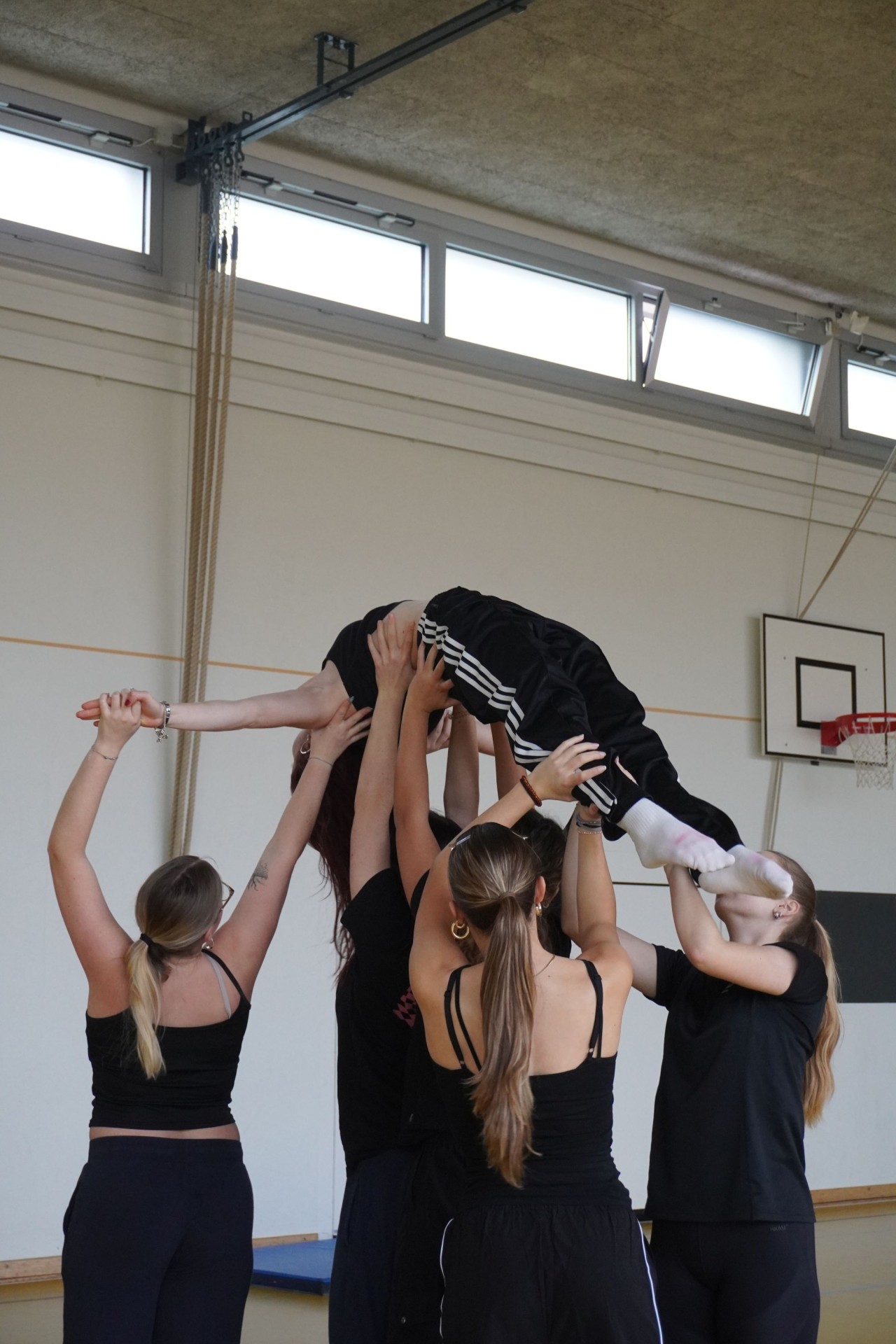 Fünf Personen in einer Turnhalle halten eine sechste Person waagerecht in Luftpose über ihre Köpfe. Eine Basketballkorbanlage hängt im Hintergrund.