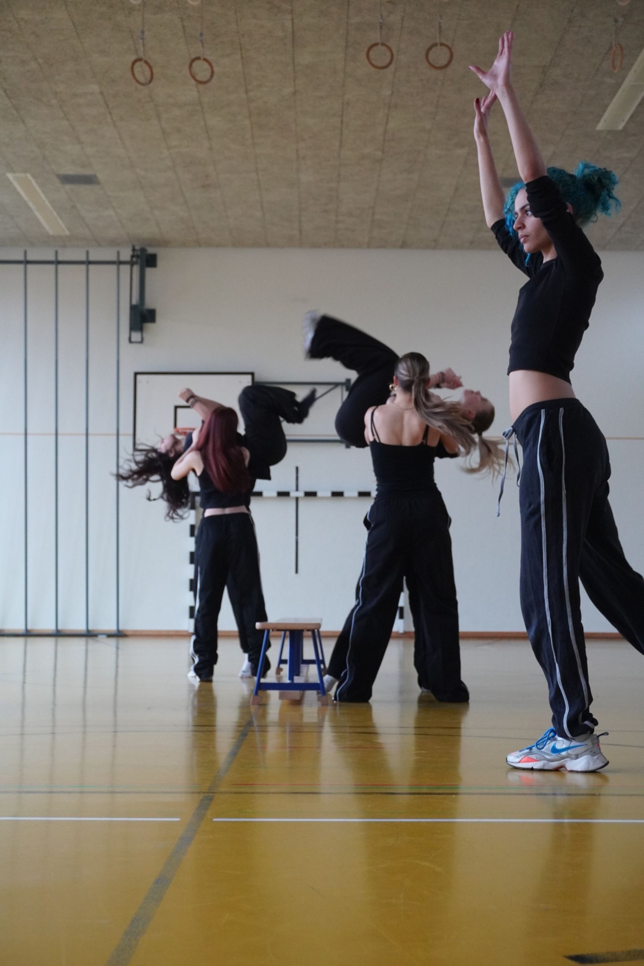 Fünf Personen führen in einer Sporthalle eine dynamische Tanz- oder Akrobatik-Choreografie aus. Eine Person wird hochgehoben, eine andere hebt die Arme, während ein kleiner Hocker in der Mitte steht.