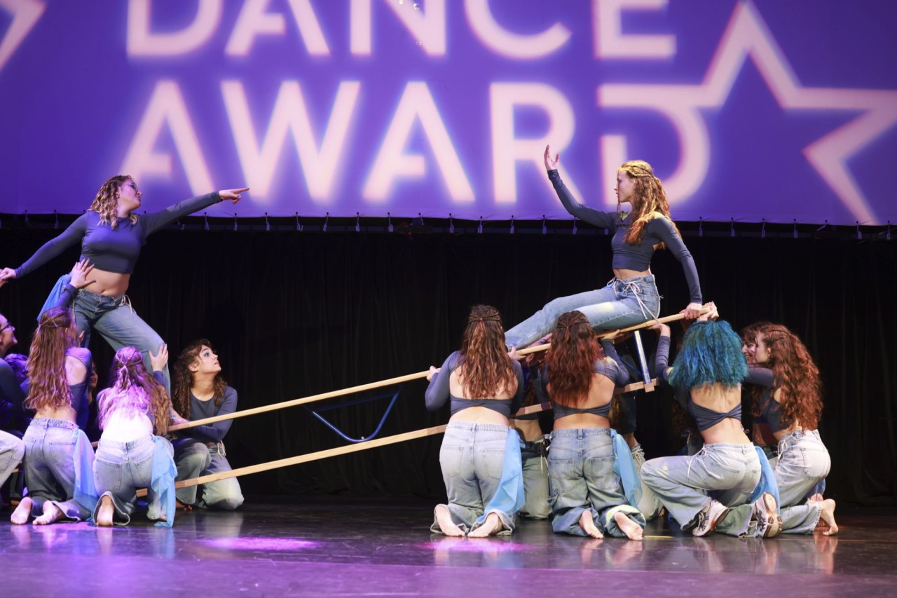Tanzgruppe in blauen Outfits führt eine synchronisierte Choreografie auf einer Bühne auf, während im Hintergrund der Schriftzug "Dance Award" und ein Sternmotiv beleuchtet sind.