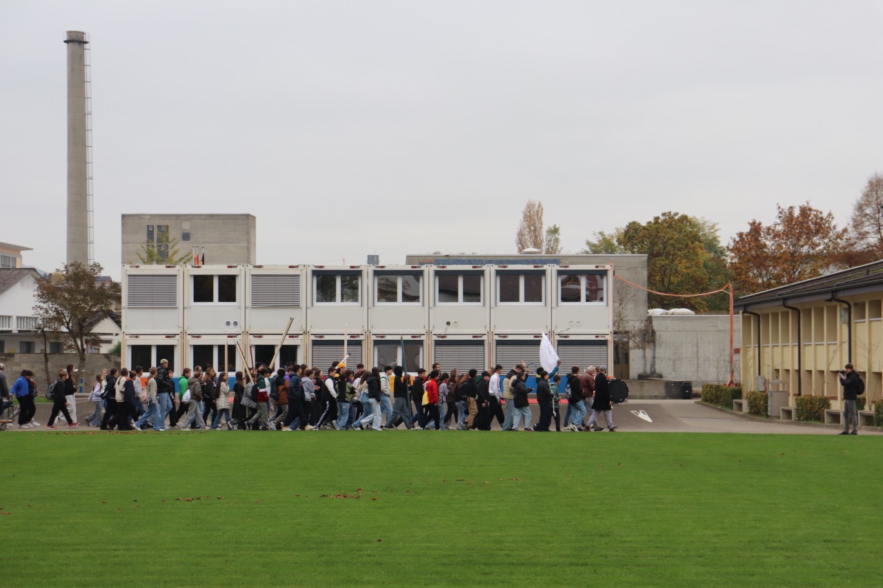 Viele junge Menschen mit Rücksäcken laufen vor einer Ansammlung gestapelter Baucontainern und tragen einen Banner.