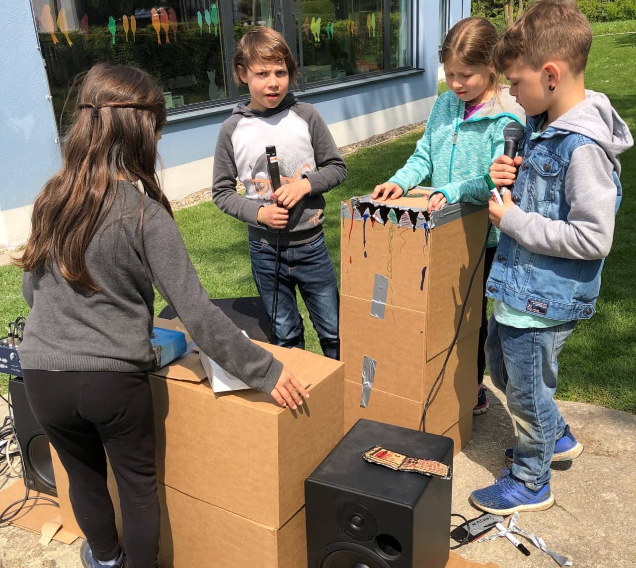 Schülerinnen und Schüler halten Mikrofone, in der Mitte stehen Kartonkisten, ein Lautsprecher steht im Vordergrund