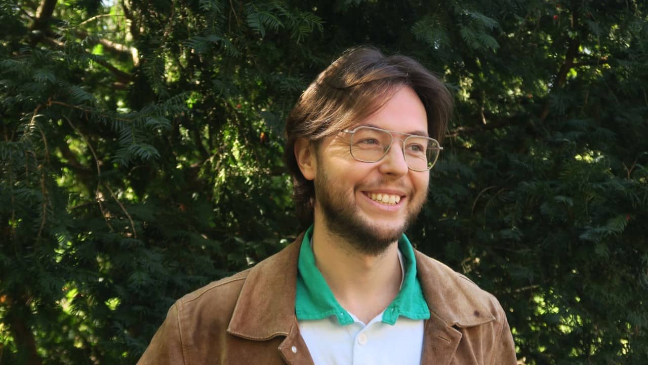 Ein lachender Mann mit Brille vor Bäumen mit grünen Blättern. 