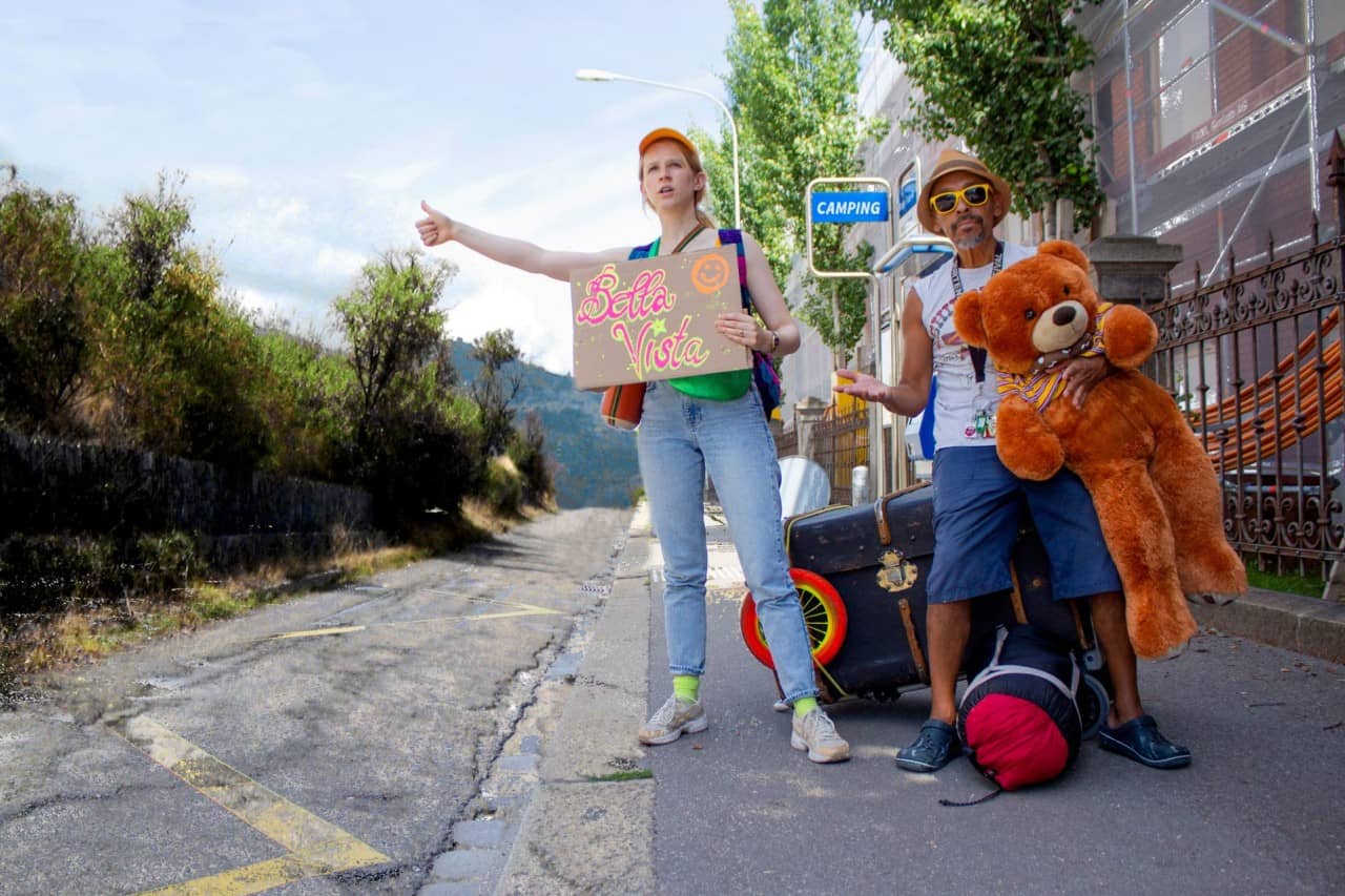 Zwei Erwachsene stehen an einer Bushaltestelle und machen Autostopp. Sie haben Gepäck und einen grossen Teddybär dabei. 