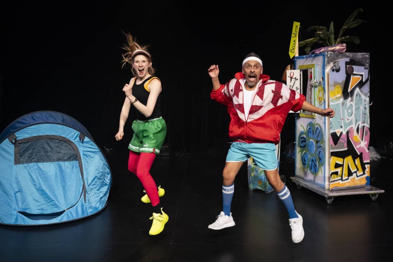 Zwei Erwachsene in Sportbekleidung springen in die Luft und schreien. Daneben steht ein Zelt und eine Box.