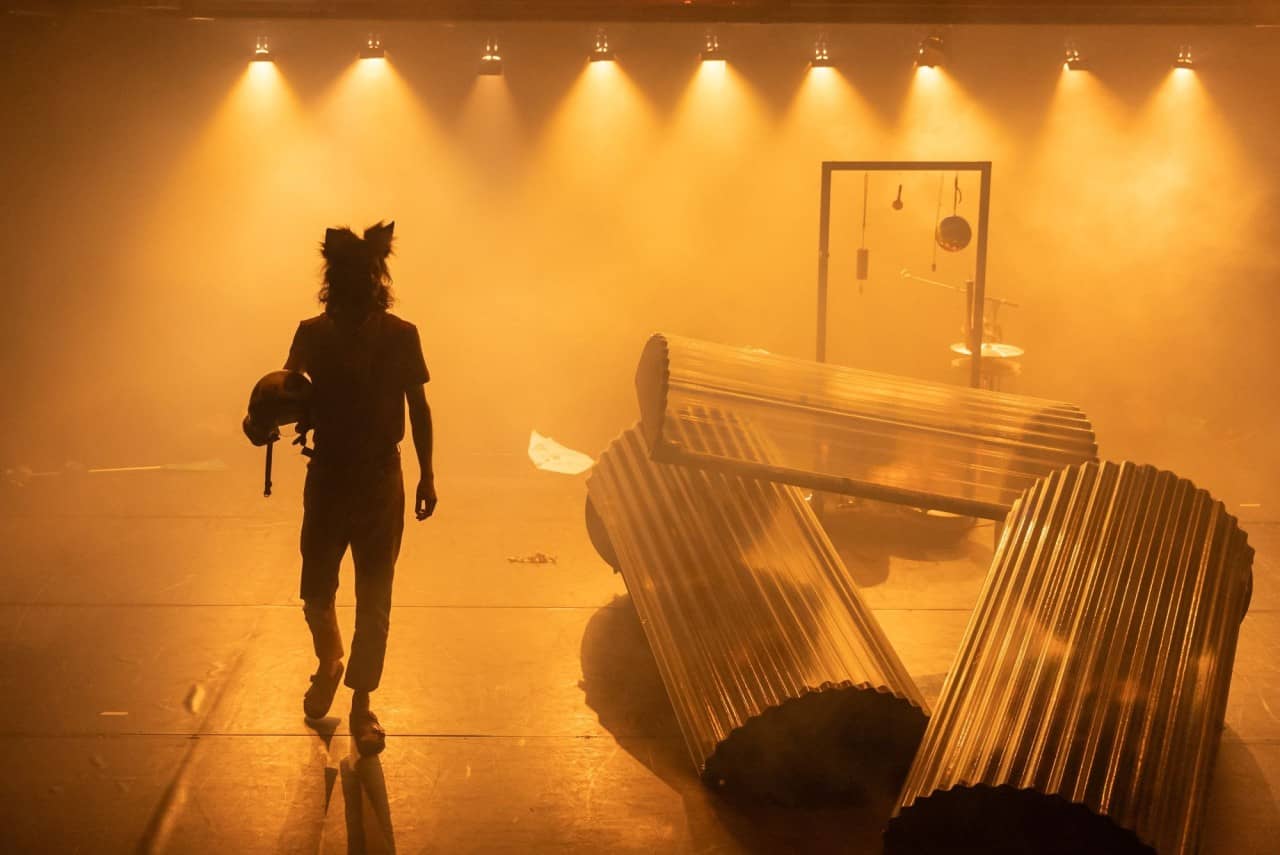 Eine als Katze verkleidete Person geht über eine von Rauch und orangem Licht durchflutete Bühne. Auf der Bühne hat es diverse Gegenstände. 