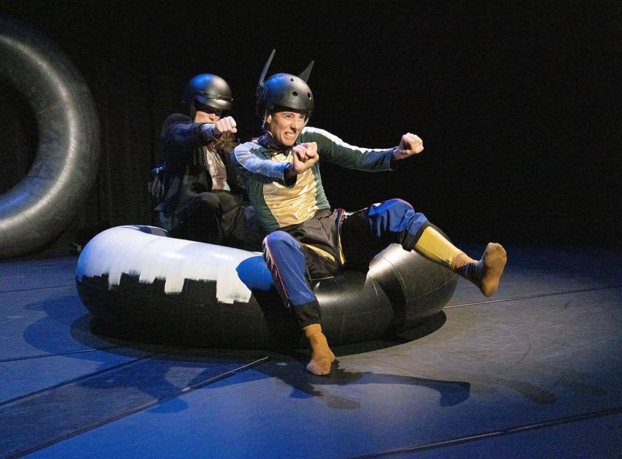 Zwei Personen mit Helm sitzen in einem aufgeblasenen Gummireifen und imitieren das Autofahren. 